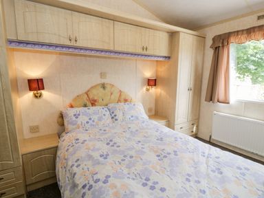 A bedroom with a double bed with floral bedding bedside cabinets built in cupboards and a window with curtains at Grange Caravan in Llangollen