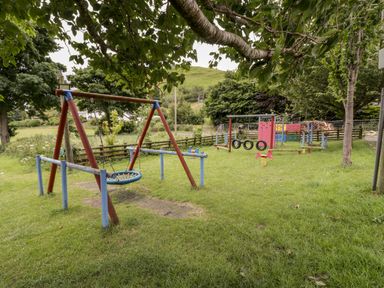 A playground with swings and climbing equipment on grass surrounded by trees at Grange Caravan in Llangollen