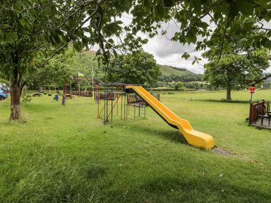 A playground with a yellow slide swings picnic tables and trees in a grassy area at Grange Caravan in Llangollen