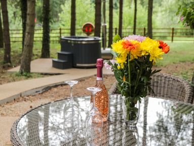 An outdoor glass table with a bottle of rose wine two inverted wine glasses and a vase of yellow orange and pink flowers with a hot tub and trees in the background at Lake View Lodge in Wykeham near East Ayton