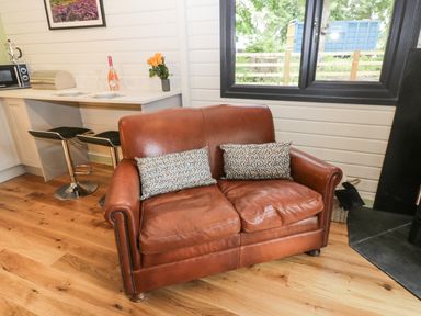 A brown leather sofa with two patterned cushions in a room with wooden floor near a kitchen counter with two black stools at Lake View Lodge in Wykeham near East Ayton