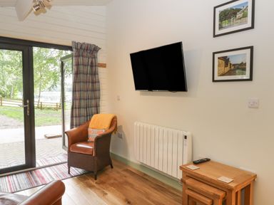 A room with a leather armchair, wall-mounted television, wooden tables, framed pictures, and glass doors leading outside at Lake View Lodge in Wykeham near East Ayton