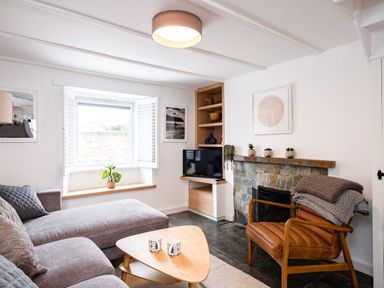 A living room with a gray sofa a wooden triangular coffee table a leather chair with blankets a stone fireplace a TV and a window at West Cottage in Rock