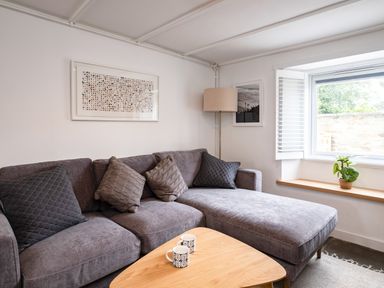 A living room with a grey sectional sofa with pillows a wooden coffee table with two mugs a floor lamp a window with shutters and a small plant on the windowsill at West Cottage in Rock