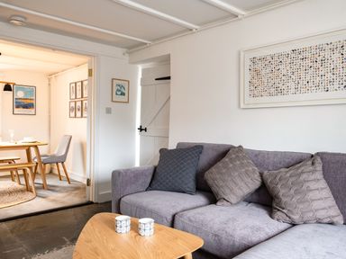 A living room with a grey sofa with cushions and a wooden coffee table with cups next to a dining area with a wooden table and chairs at West Cottage in Rock