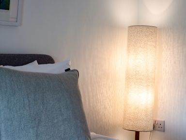 A bedroom corner with a bed with pillows and a lit cylindrical floor lamp at West Cottage in Rock