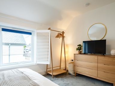 A bedroom with a bed a window an open shutter a wooden clothes rack with hangers a wooden dresser a television a round wall mirror a lamp and a plant at West Cottage in Rock