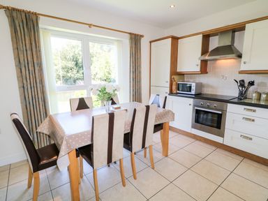 A kitchen with a dining table and chairs next to a window with curtains and cabinets with appliances at 4 The Cloisters in Bushmills County Antrim