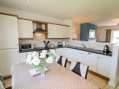 A kitchen with white cabinets a dining table with flowers and a living area visible at 4 The Cloisters Bushmills County Antrim