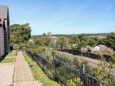 A paved walkway with grass and metal fence beside houses and trees and a church tower in the distance at 4 The Cloisters in Bushmills County Antrim