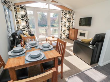 A dining area with a wooden table set for four and a living room with black leather sofas near large windows at Pinecone Lodge - No 9 Felmoor Park near Felton