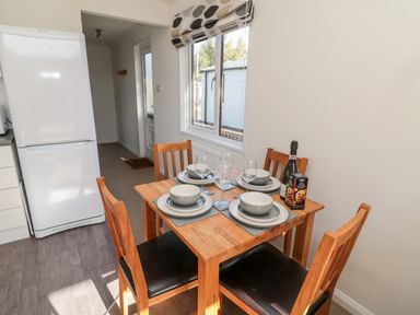 A dining area with a wooden table set for four near a window and a white refrigerator in the kitchen at Pinecone Lodge - No 9 Felmoor Park near Felton
