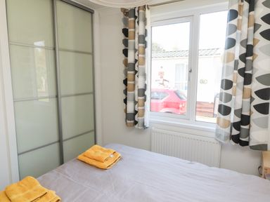 A bedroom with a bed towels on it a window with patterned curtains and a wardrobe with frosted glass doors at Pinecone Lodge - No 9 Felmoor Park near Felton