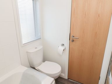 A bathroom with a toilet next to a wooden door and a window with blinds at Pinecone Lodge - No 9 Felmoor Park near Felton
