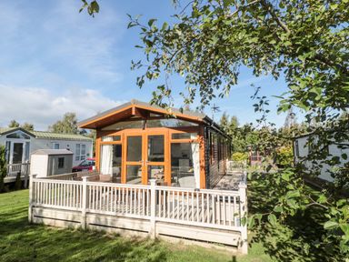 A wooden lodge with a deck surrounded by a white railing in a grassy area at Pinecone Lodge - No 9 Felmoor Park near Felton