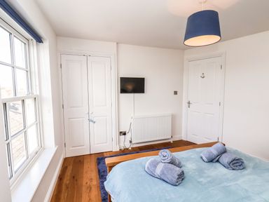 A bedroom with a wooden floor a double bed with blue bedding rolled towels on the bed a blue rug a white wardrobe a wall-mounted TV and white doors at Navigators Rest Whitby