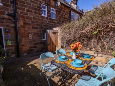 An outdoor patio with a black metal table set with orange placemats blue plates and bowls four blue metal chairs and a vase with orange flowers at Navigators Rest in Whitby