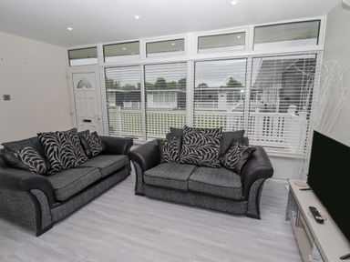 A living room with two sofas, a television on a stand, and windows with blinds at 19 Cherry Park in Chapel St Leonards