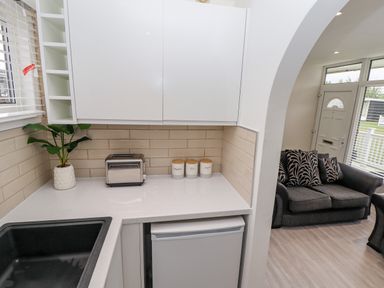 A kitchen corner with white cabinets a toaster three labeled jars a plant and a black sink next to a living room with a gray sofa and patterned cushions at 19 Cherry Park in Chapel St Leonards