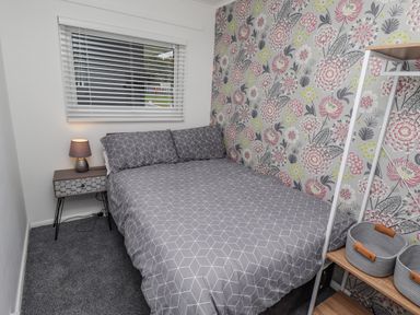 A bedroom with a bed with patterned bedding a bedside table with a lamp a window with blinds and shelving with baskets at 19 Cherry Park in Chapel St Leonards