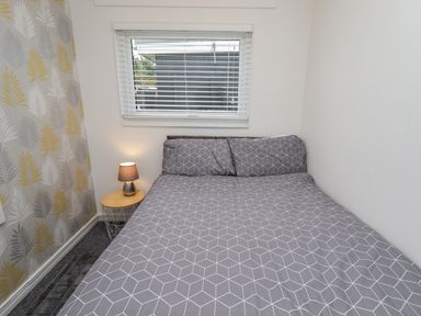 A bedroom with a double bed gray patterned bedding a small round wooden bedside table with a lamp and patterned wallpaper at 19 Cherry Park in Chapel St Leonards