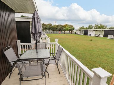 A balcony with a table umbrella and four chairs overlooking a grassy lawn and black and white buildings at 19 Cherry Park in Chapel St Leonards
