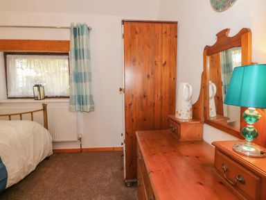 A bedroom with a bed next to a window with curtains a wooden wardrobe a wooden dresser with a mirror a green lamp and a ceramic pitcher at Burnt Oak Stables in Mynydd Isa