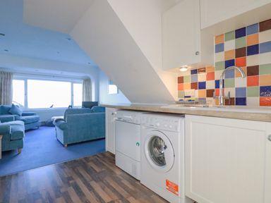 A kitchen area with washing machine dishwasher and colorful tiled backsplash next to a living room with blue sofas and large window at Sea View in Mevagissey