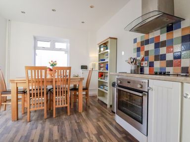A kitchen with a wooden dining table and chairs a bookshelf and a stove with a colorful tiled backsplash at Sea View in Mevagissey