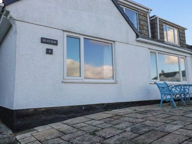 Exterior view of a white house with windows and blue metal bench and table on a paved patio at Sea View in Mevagissey