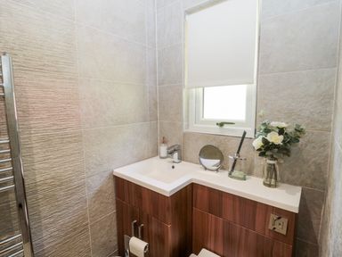A bathroom with a corner sink and wooden cabinets below a window with a blind at Lodge 32 in Otterburn