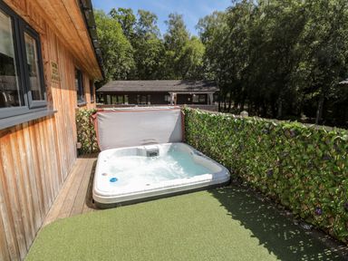 An outdoor hot tub on a deck next to a wooden building with a leafy fence and trees at Lodge 32 in Otterburn