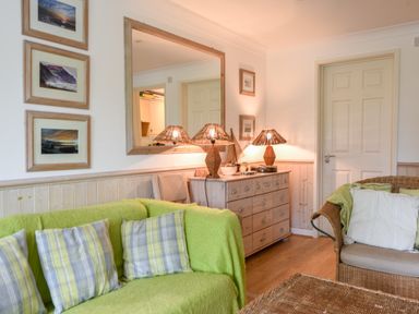 A living room with green sofa cushions a wicker chair lamps on a wooden sideboard and framed pictures on the wall at 52 Fernhill Heights in Charmouth