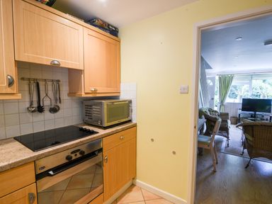 A kitchen with wooden cabinets and a microwave next to a living room with wicker chairs and a television at 52 Fernhill Heights in Charmouth