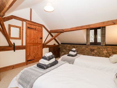 A bedroom with two single beds with folded towels on top and a wooden door with exposed beams and a window with plaid curtains at Bullfinch Cottage in Kingsland