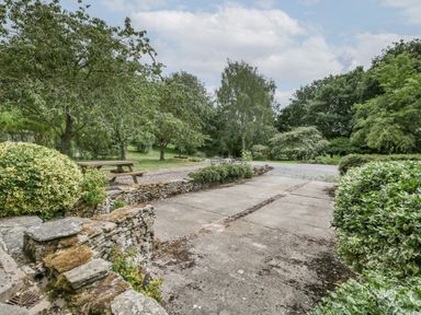 An outdoor area with a concrete driveway surrounded by trees bushes and stone walls at Bullfinch Cottage in Kingsland