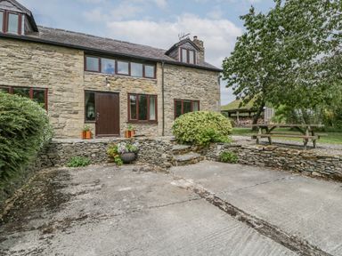 A stone house with a brown door and windows surrounded by stone walls bushes a wooden picnic table and a tree at Bullfinch Cottage in Kingsland