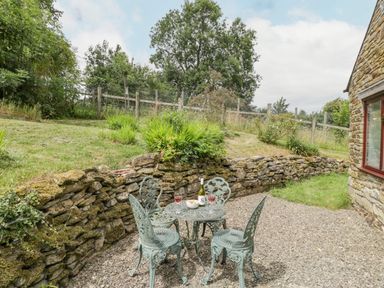 A patio area with metal table and four chairs and a bottle with two glasses and bowl against a stone wall at Fieldfare in Kingsland