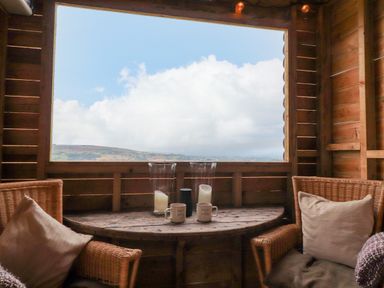 A wooden room with two wicker chairs a half round wooden table with candles and mugs and a large window showing a cloudy sky at Heather Hut at Copy House Hideaway in Earby