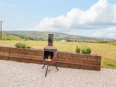 An outdoor fire pit with wood on gravel in front of a wooden planter and a grassy field at Heather Hut at Copy House Hideaway in Earby