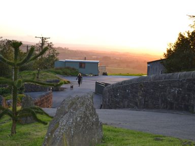 A person walking a dog on a paved path near a stone wall with a small building and hills in the background at Heather Hut at Copy House Hideaway in Earby