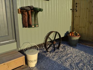 A wall with hanging boots a kindling bucket a metal wheel and a pot with firewood on gravel outside a building at Heather Hut at Copy House Hideaway in Earby