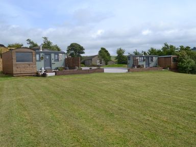 A large grassy area with two small cabin-like structures and wooden sheds in a rural setting at Heather Hut at Copy House Hideaway in Earby