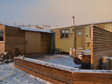 A small wooden cabin with a fenced outdoor seating area and shed in a snowy yard with sheep in the background at Heather Hut at Copy House Hideaway in Earby