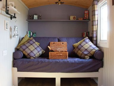 A small seating area with a purple cushioned bench couch plaid pillows a wicker basket with a bottle and glasses books and board games on shelves at Heather Hut at Copy House Hideaway in Earby