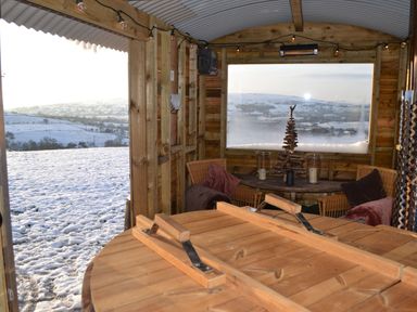 An interior wooden seating area with round table cushions and a window view of snowy hills at Heather Hut at Copy House Hideaway in Earby