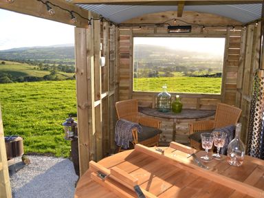 A wooden outdoor shelter with wicker chairs a small table bottles and glasses with green fields visible outside Heather Hut at Copy House Hideaway Earby