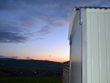 A metal shed with a wooden fence in front of a scenic hillside at dusk at Heather Hut at Copy House Hideaway in Earby