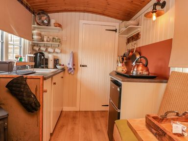A small kitchen with wooden ceiling and floor with shelves holding cups and bowls and a kettle on the counter at Heather Hut at Copy House Hideaway in Earby