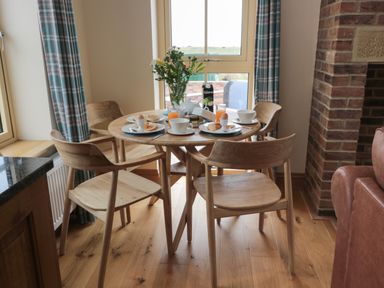 A round wooden dining table set for breakfast with four wooden chairs in a room with a window and brick fireplace at Whinny Cottage in Embleton